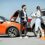 Coppia discute dopo un incidente neopatentato. Auto arancione con cofano aperto e cono arancione.