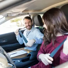 Istruttore sorridente allaccia la cintura per l'esame pratico patente B motorizzazione. La candidata si prepara.