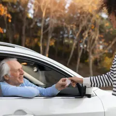 Un uomo anziano in auto sorride mentre riceve documenti per rinnovo patente dopo 70 anni da una giovane donna.