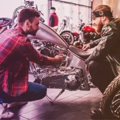 Due uomini esaminano un motore di moto custom. Uno chiede quanto costa targare una moto, mentre l'altro mostra i dettagli.