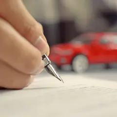Mano che firma un foglio complementare con una penna stilografica, con una macchinina rossa sfocata sullo sfondo.