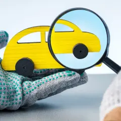 Mano guantata esamina un'auto giocattolo gialla con una lente d'ingrandimento, suggerendo un'indagine su una possibile truffa assicurazioni.