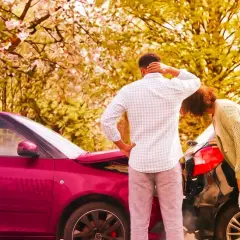 Coppia esamina i danni dopo un incidente. Il cofano di un'auto rossa è aperto, mentre un'altra auto nera è danneggiata. Il **RCA significato** è la copertura assicurativa obbligatoria.