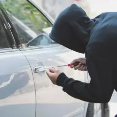 Persona con cappuccio tenta di scassinare la portiera di un'auto, un'azione che potrebbe essere coperta dalla polizza assicurativa contro il rischio di furto o incendio.