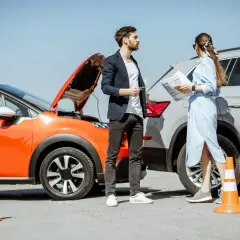 Coppia discute dopo un incidente neopatentato. Auto arancione con cofano aperto e cono arancione.