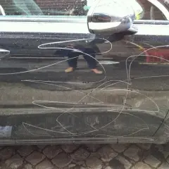 Graffi bianchi sulla portiera di un'auto scura, segno di denuncia atti vandalici auto. Riflesso di una persona con pantaloni scuri e scarpe chiare.