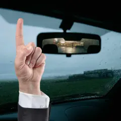 Mano con dito puntato in alto, ricorda che il bollino revisione auto va esposto. Vista dall'interno di un'auto con pioggia sul parabrezza.