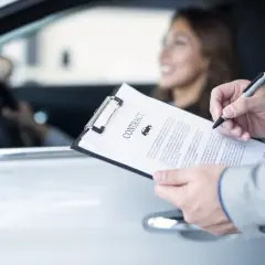 Uomo in giacca firma un contratto d'assicurazione auto. La donna al volante sorride. La decorrenza assicurazione è un dettaglio importante.