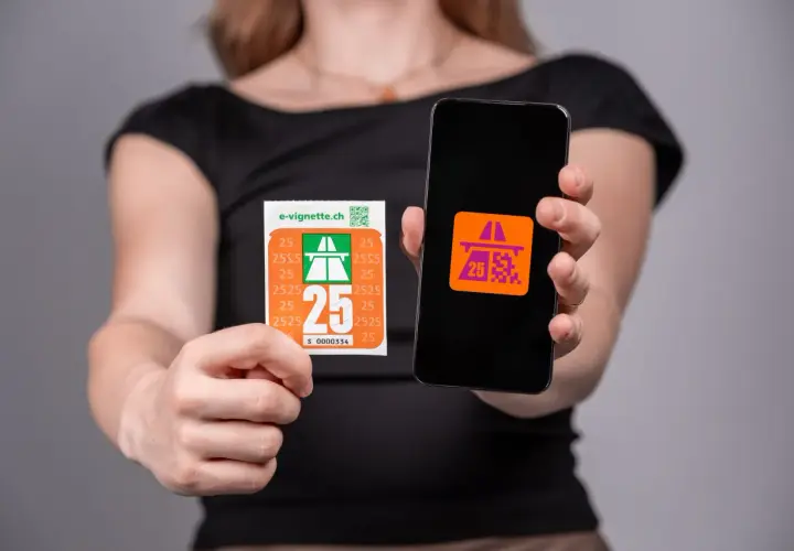 Una persona mostra una vignetta autostradale e uno smartphone con un'icona simile. Per andare a Lugano serve la vignetta.