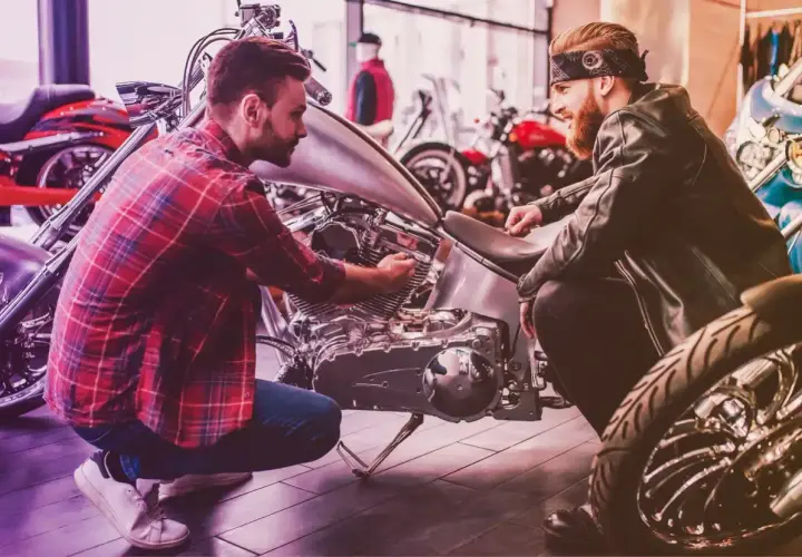 Due uomini esaminano un motore di moto custom. Uno chiede quanto costa targare una moto, mentre l'altro mostra i dettagli.