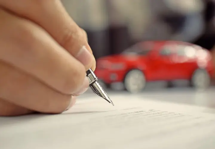 Mano che firma un foglio complementare con una penna stilografica, con una macchinina rossa sfocata sullo sfondo.