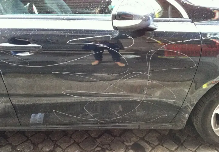 Graffi bianchi sulla portiera di un'auto scura, segno di denuncia atti vandalici auto. Riflesso di una persona con pantaloni scuri e scarpe chiare.
