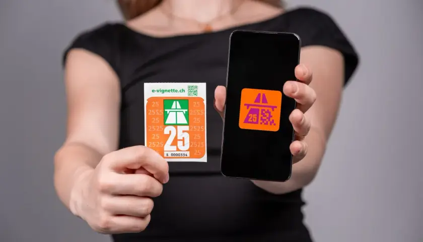 Una persona mostra una vignetta autostradale e uno smartphone con un'icona simile. Per andare a Lugano serve la vignetta.