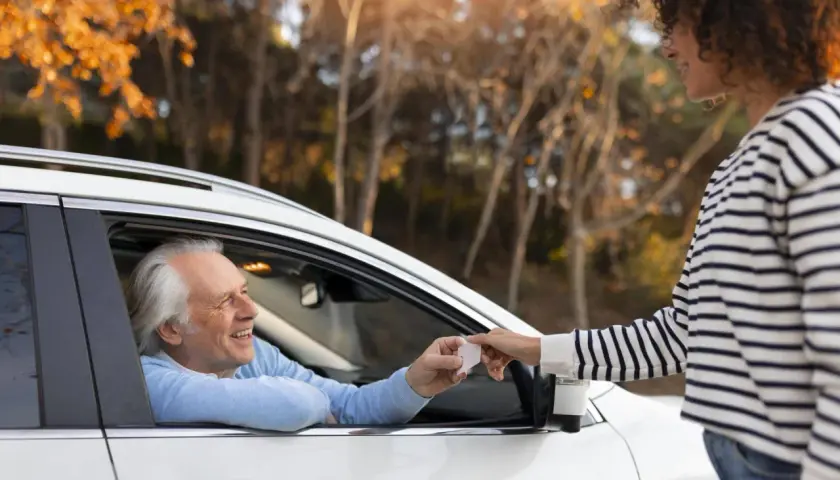 Un uomo anziano in auto sorride mentre riceve documenti per rinnovo patente dopo 70 anni da una giovane donna.
