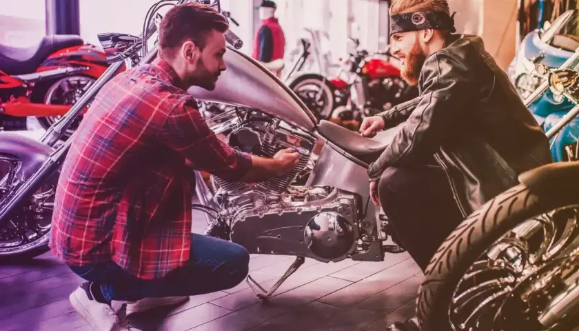 Due uomini esaminano un motore di moto custom. Uno chiede quanto costa targare una moto, mentre l'altro mostra i dettagli.