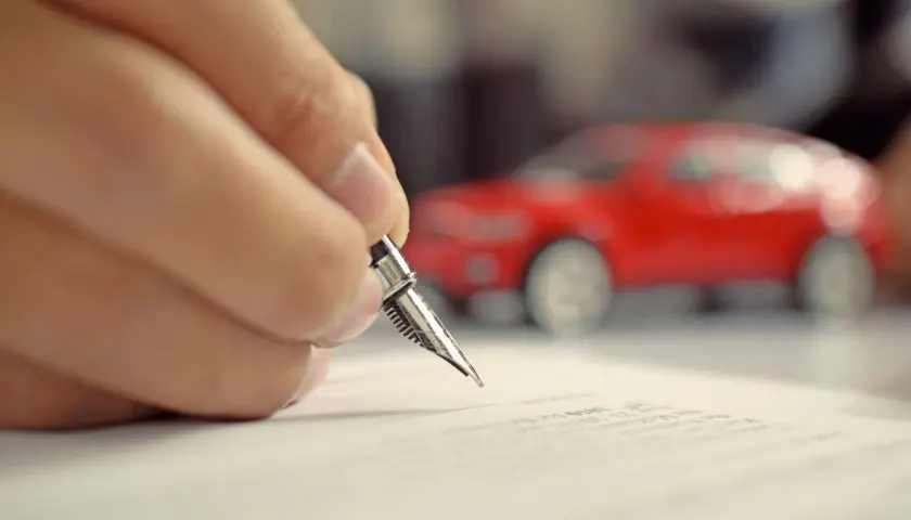 Mano che firma un foglio complementare con una penna stilografica, con una macchinina rossa sfocata sullo sfondo.