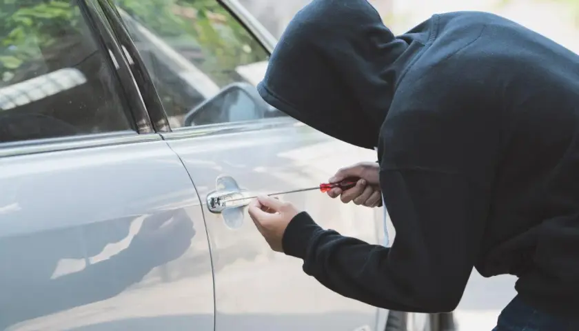 Persona con cappuccio tenta di scassinare la portiera di un'auto, un'azione che potrebbe essere coperta dalla polizza assicurativa contro il rischio di furto o incendio.