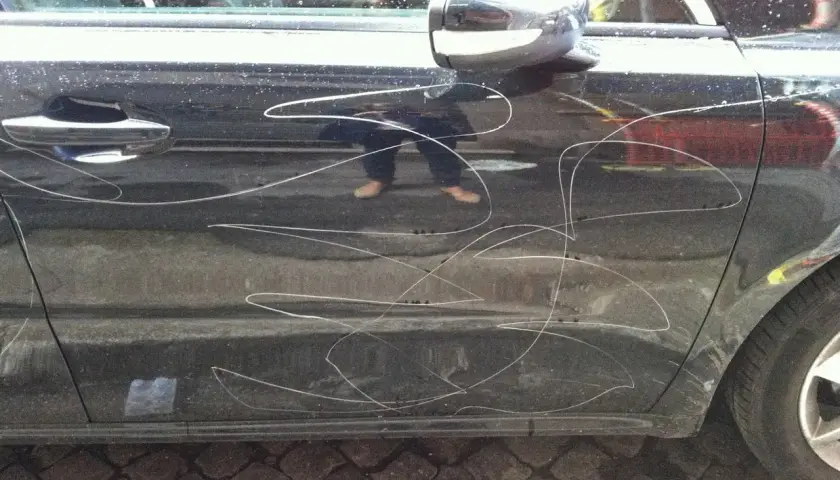 Graffi bianchi sulla portiera di un'auto scura, segno di denuncia atti vandalici auto. Riflesso di una persona con pantaloni scuri e scarpe chiare.
