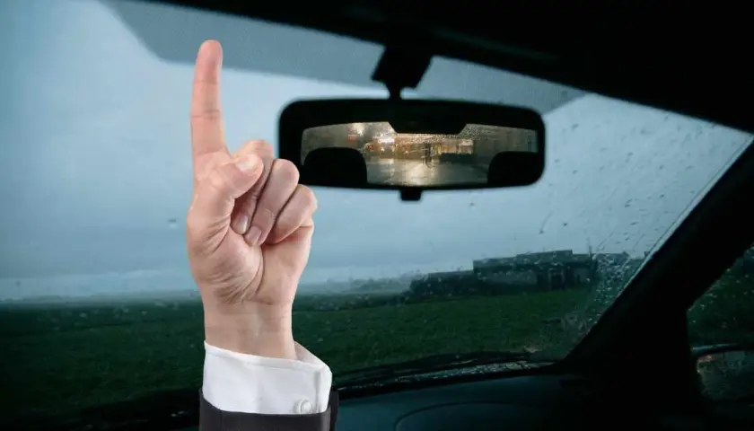Mano con dito puntato in alto, ricorda che il bollino revisione auto va esposto. Vista dall'interno di un'auto con pioggia sul parabrezza.