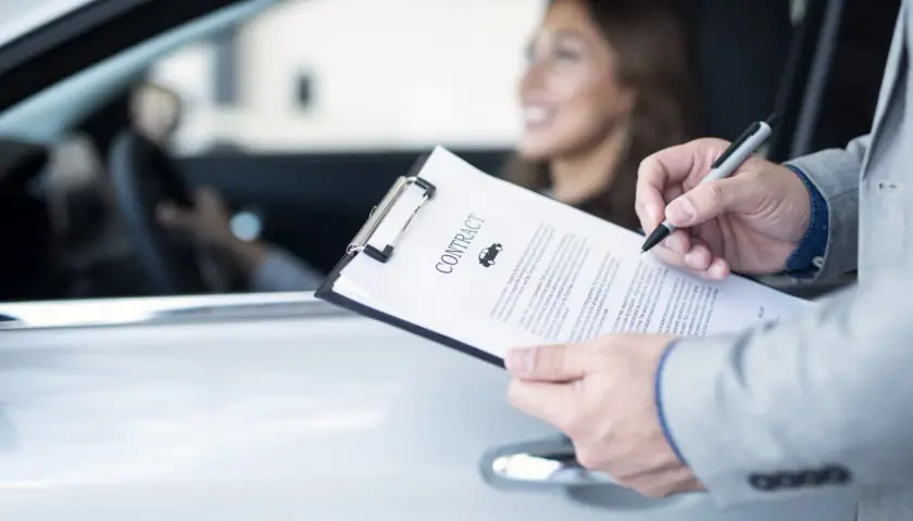 Uomo in giacca firma un contratto d'assicurazione auto. La donna al volante sorride. La decorrenza assicurazione è un dettaglio importante.