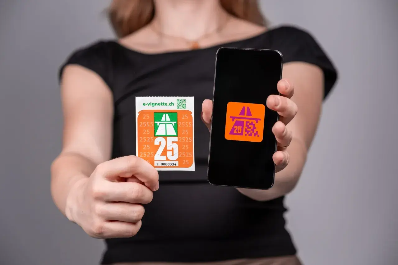 Una persona mostra una vignetta autostradale e uno smartphone con un'icona simile. Per andare a Lugano serve la vignetta.