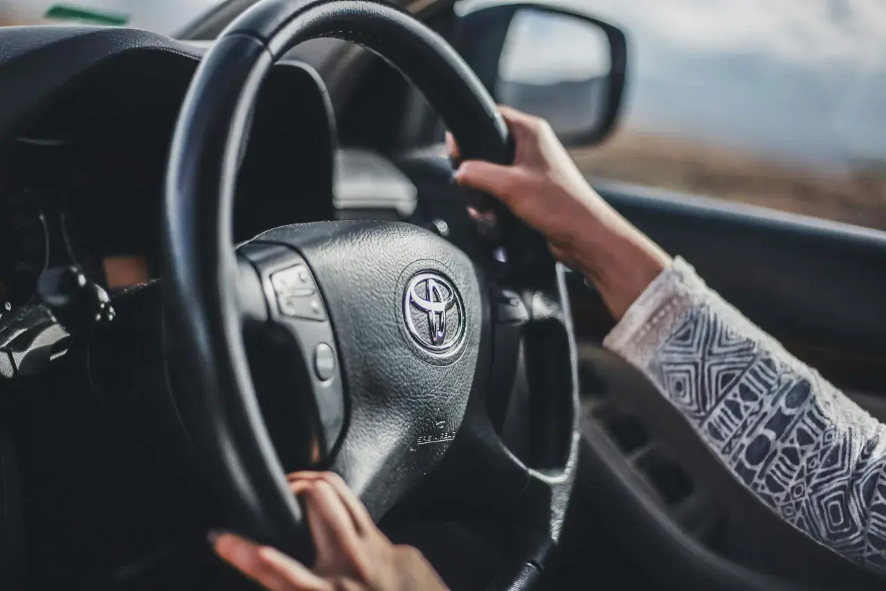 Mani sul volante di una Toyota, con un braccio che indossa una maglia decorata. Finalmente posso guidare una macchina non intestata a me, verso l'avventura.