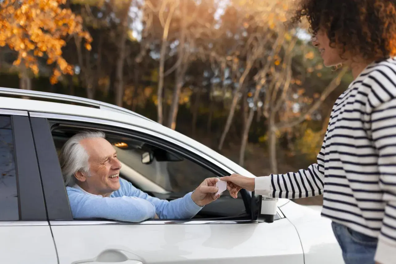 Un uomo anziano in auto sorride mentre riceve documenti per rinnovo patente dopo 70 anni da una giovane donna.