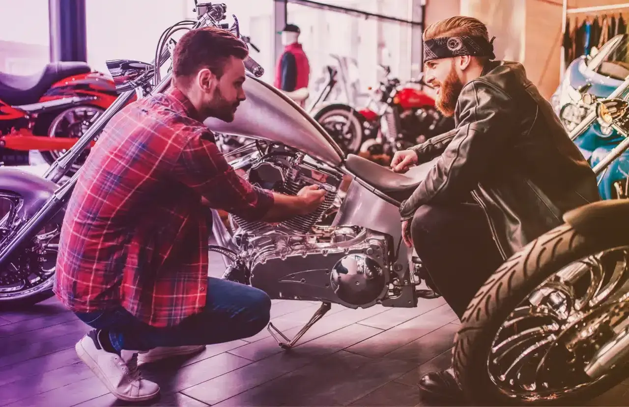 Due uomini esaminano un motore di moto custom. Uno chiede quanto costa targare una moto, mentre l'altro mostra i dettagli.