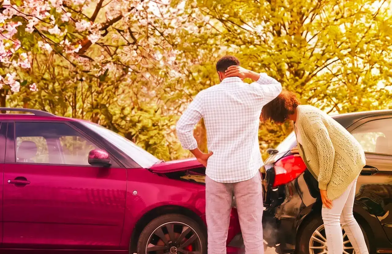 Coppia esamina i danni dopo un incidente. Il cofano di un'auto rossa è aperto, mentre un'altra auto nera è danneggiata. Il **RCA significato** è la copertura assicurativa obbligatoria.