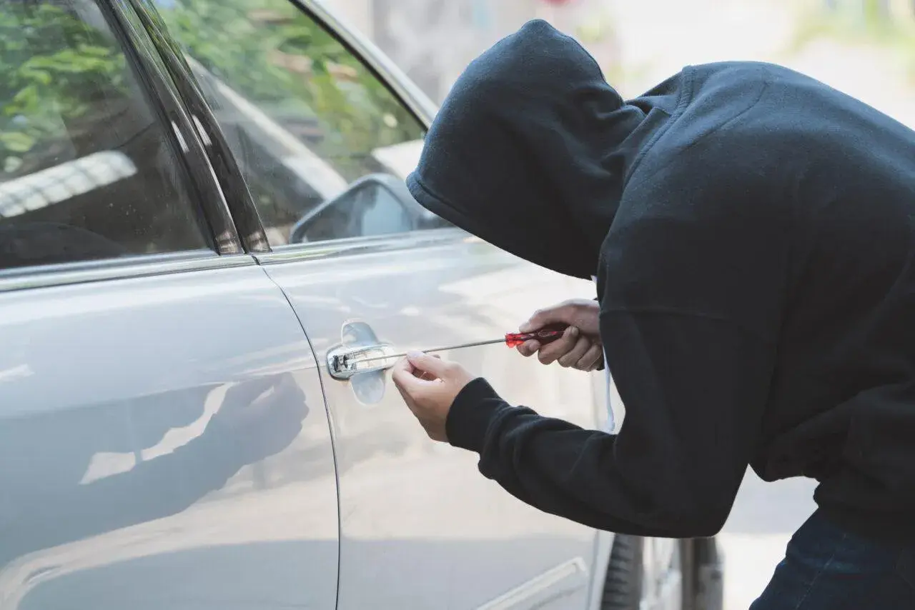 Persona con cappuccio tenta di scassinare la portiera di un'auto, un'azione che potrebbe essere coperta dalla polizza assicurativa contro il rischio di furto o incendio.