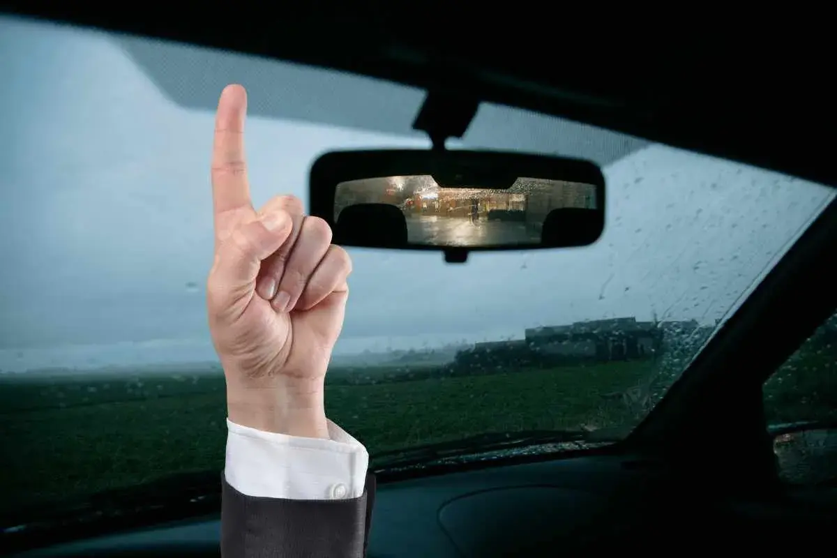 Mano con dito puntato in alto, ricorda che il bollino revisione auto va esposto. Vista dall'interno di un'auto con pioggia sul parabrezza.