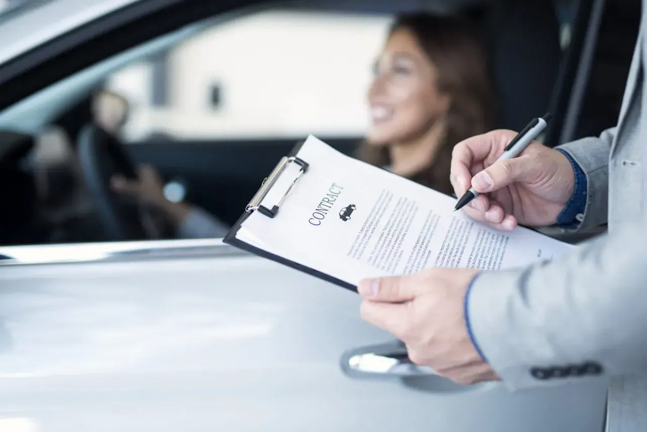 Uomo in giacca firma un contratto d'assicurazione auto. La donna al volante sorride. La decorrenza assicurazione è un dettaglio importante.