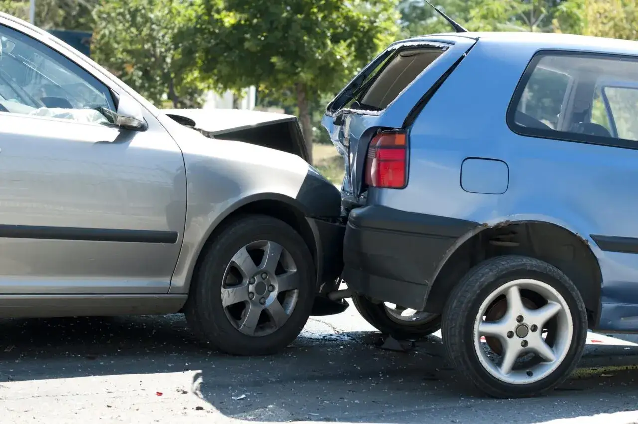 Due auto danneggiate dopo un incidente. Il cofano di un'auto grigia è sollevato, mentre un'auto blu ha il lunotto posteriore rotto. Un classico incidente neopatentato.
