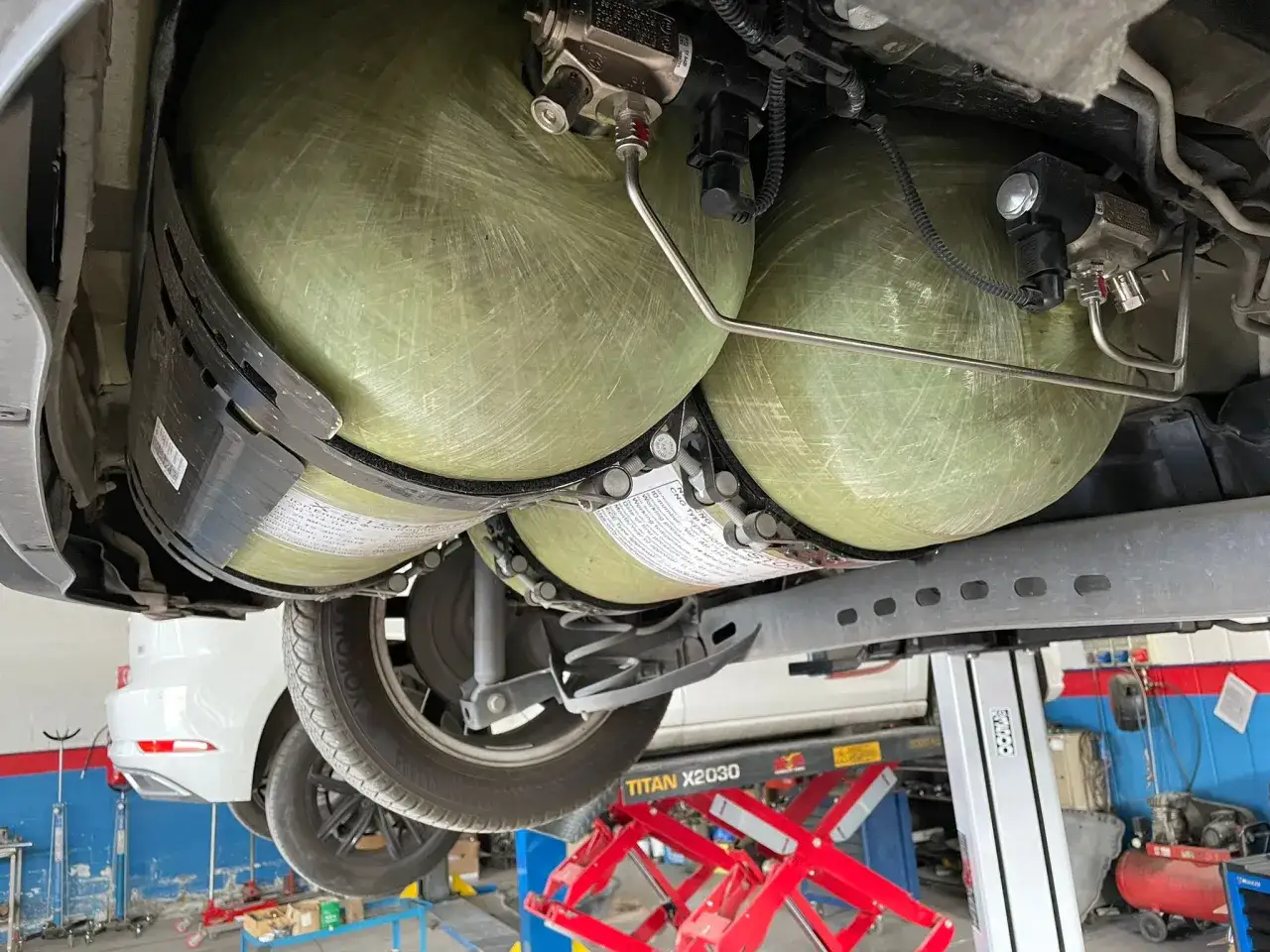 Sotto un'auto in officina, si vedono le bombole metano revisione, pronte per essere controllate.