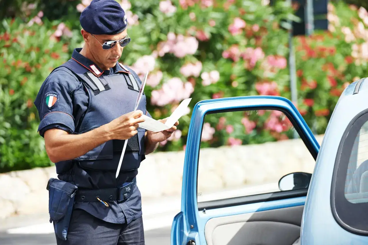 Agente di polizia italiano con berretto blu e occhiali da sole controlla documenti vicino a un'auto azzurra. Forse sta verificando se posso guidare una macchina non intestata a me.