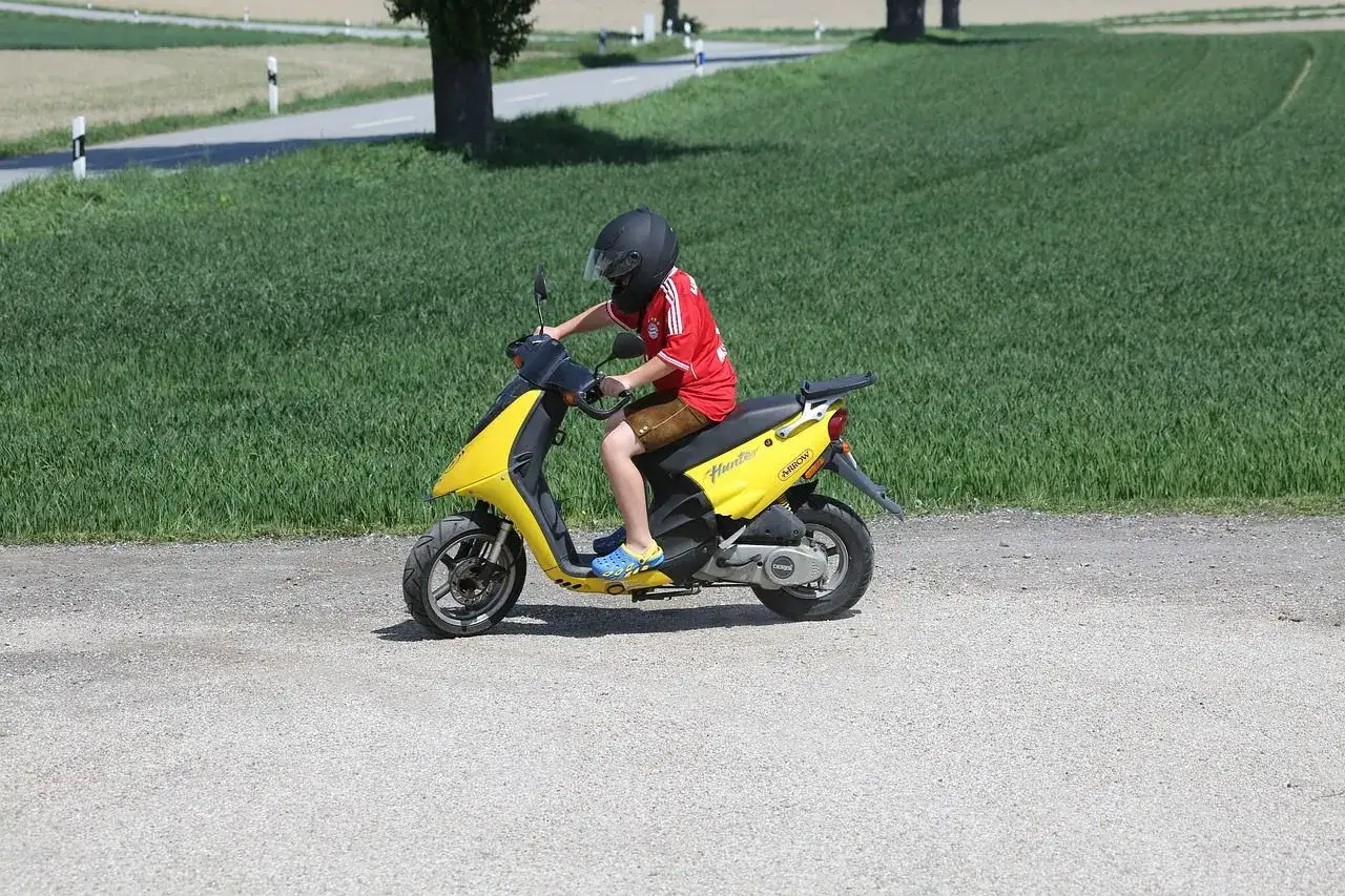 Ragazzo con casco guida un motorino giallo su una strada sterrata, felice di poter guidare da solo con il foglio rosa.